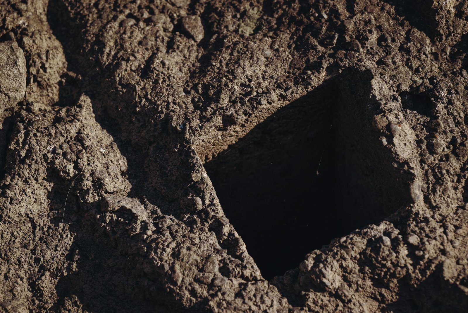 Close-up view of a rectangular hole dug into a rough, textured soil surface, showcasing geological features.